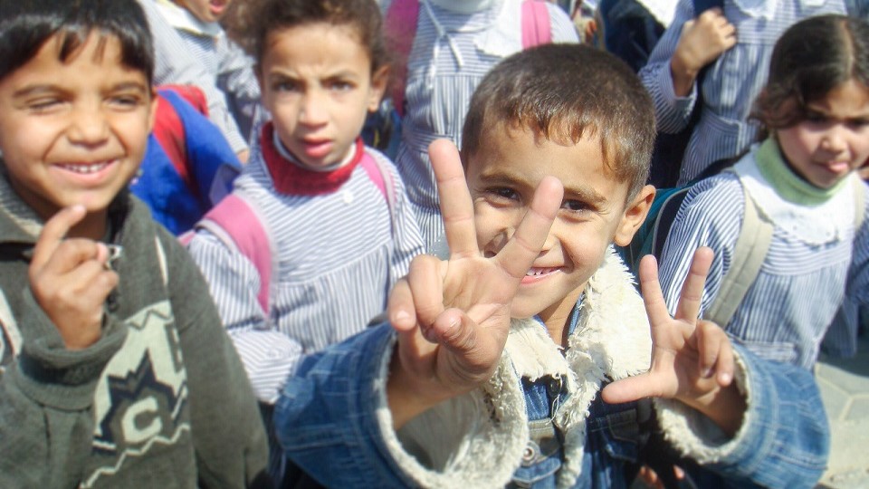 Smiling Gazan schoolchild holding up hands showing five fingers in a crowd of other children