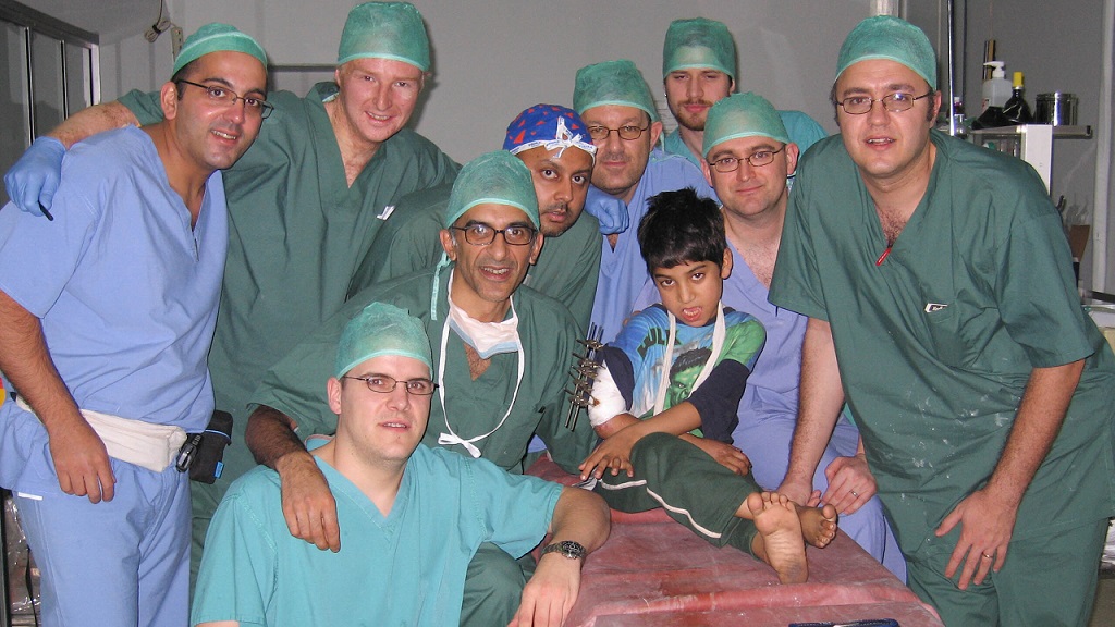 Group of Nine surgical MIST members in scrubs with young boy with right arm in external fixator sitting on a theatre table at Rawalpindi eye hospital in Pakistan 2005